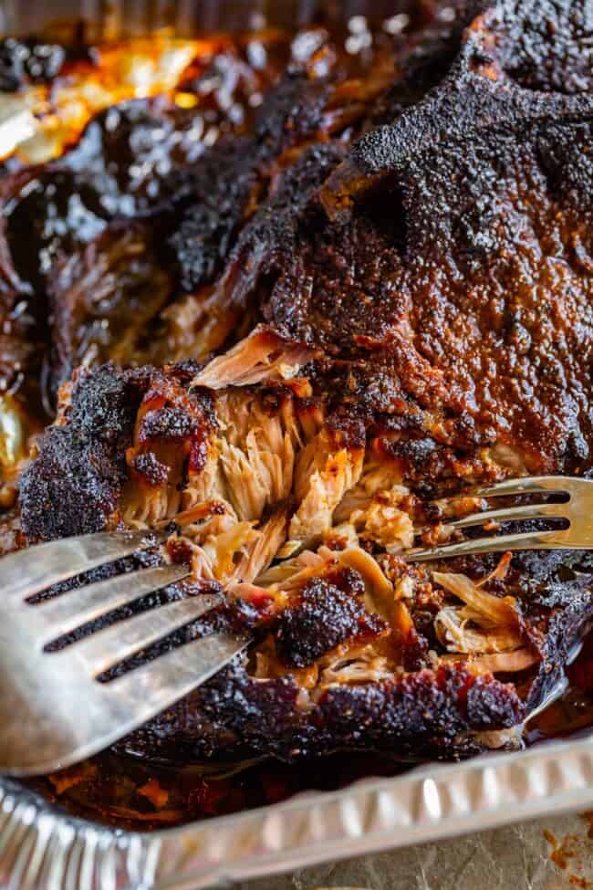 two forks shredding smoked pulled pork with blackened bark edge in an aluminum pan.