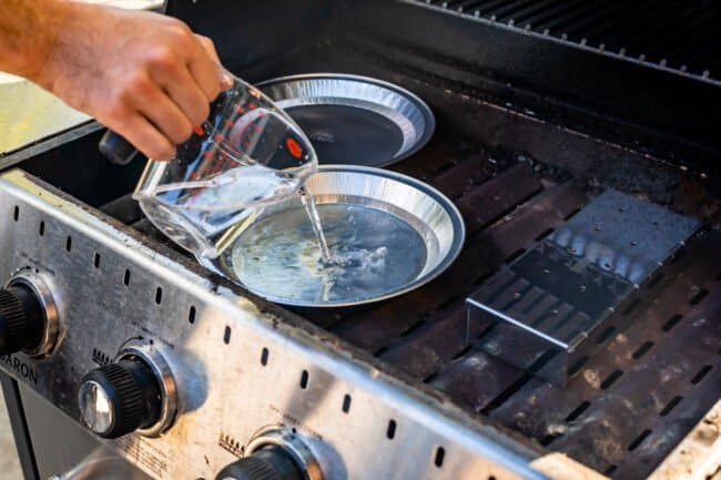 pouring water into a tin pie pan set on the heating plates of an outdoor grill.