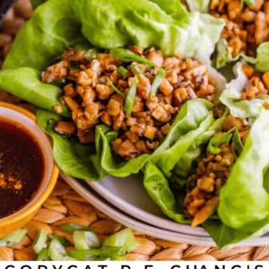 Close-up of chicken lettuce wraps filled with diced chicken and vegetables on fresh lettuce leaves, served with a small bowl of dipping sauce. Text reads Copycat P.F. Changs Chicken Lettuce Wraps - The Food Charlatan.