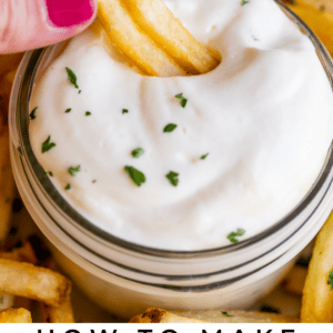 A hand with pink nail polish dips a French fry into a jar of creamy garlic aioli, surrounded by more fries. Text at the bottom reads: Easy Garlic Aioli. The Food Charlatan.
