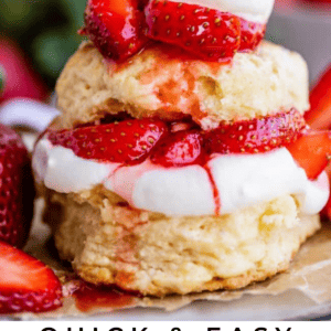 A close-up of a delicious strawberry shortcake dessert topped with fresh strawberries and whipped cream, stacked in two layers, with whole strawberries in the background and text that reads Quick & Easy Strawberry Shortcake.