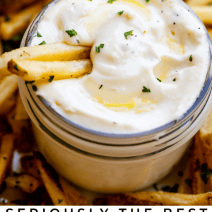 A mason jar filled with creamy garlic aioli, topped with chopped parsley, sits among golden French fries. Two fries are dipped in the aioli. Text below reads: Seriously classic garlic aioli. The Food Charlatan.