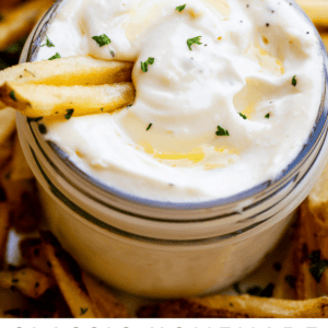 A jar of creamy garlic aioli topped with parsley sits on a plate of French fries, with two fries dipped in the tangy dipping sauce. Text below reads Classic Garlic Aioli with Cheater Version - The Food Charlatan.