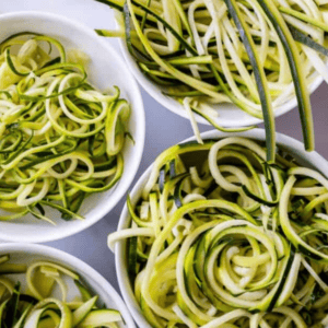 Five white bowls filled with spiralized and ribboned zucchini noodles are arranged on a gray surface. Text above reads, How to Make Zucchini Noodles—discover how to make zoodles using fresh zucchini for a healthy twist.