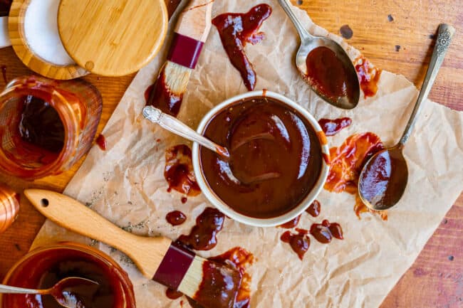 overhead shot of a bowl of bbq sauce with spoons, brushes, and parchment paper on a wooden table.