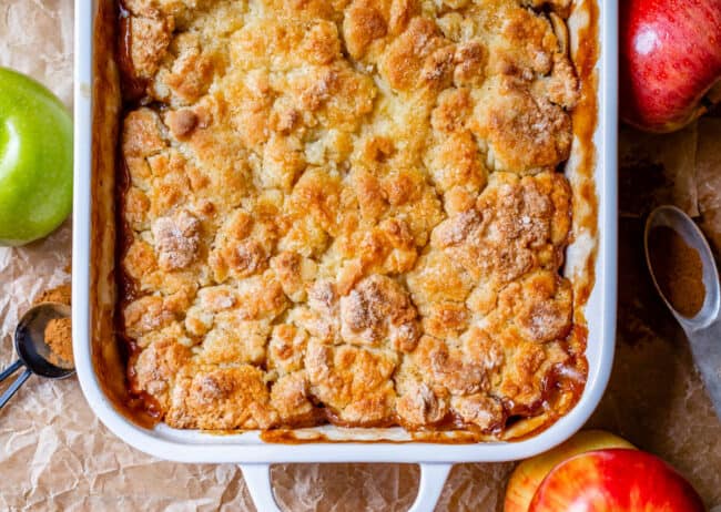 overhead shot of golden brown crisp apple cobbler in a white casserole dish with apples around it.