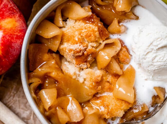 apple cobbler in a bowl with ice cream and a fork, red apple next to the bowl.