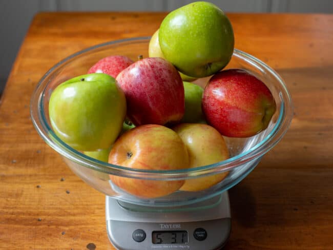 variety of apples in a clear glass bowl, sitting on a scale weighing 5 pounds.