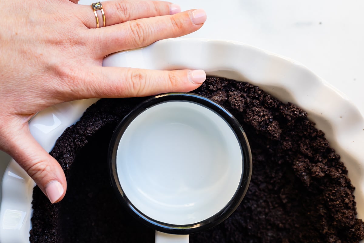 hand and measuring cup pressing oreo crumbs into a white pie dish.