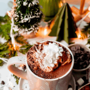 A festive mug of hot chocolate topped with whipped cream and chocolate shavings, surrounded by marshmallows, chocolate chips, and holiday decorations. Text reads Hot Chocolate.