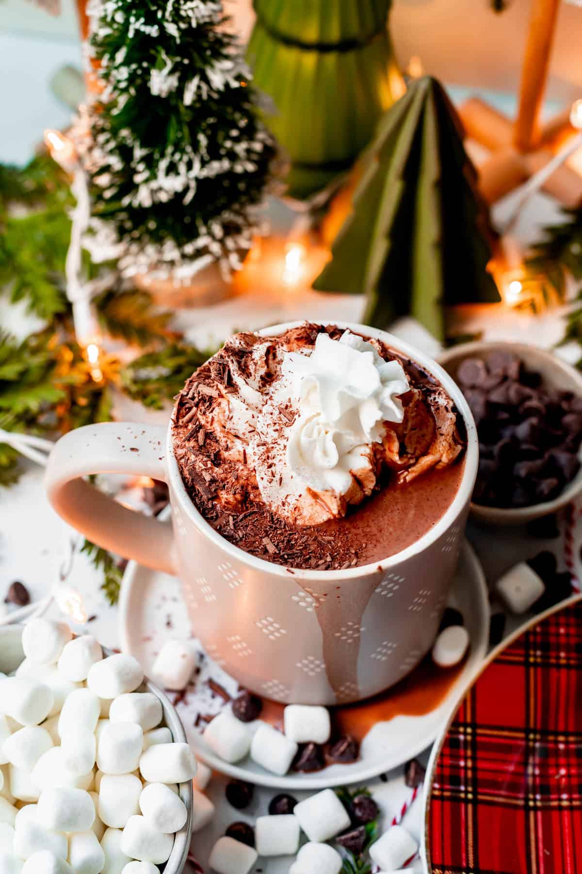 a mug of homemade hot chocolate on a table with marshmallows, chocolate chips, and mini Christmas trees.