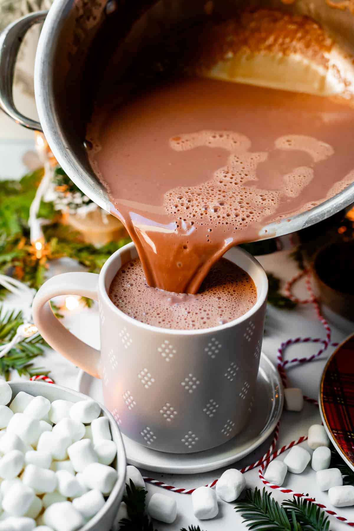 pouring hot chocolate from a pot into a gray and white mug with mini marshmallows on the table around it.