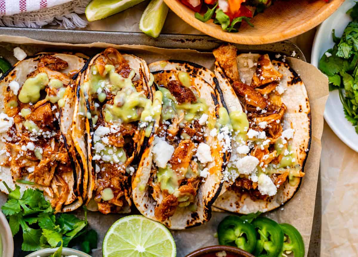 four crock pot chicken tacos side by side in charred corn tortillas with juicy meat, cotija, and green salsa.