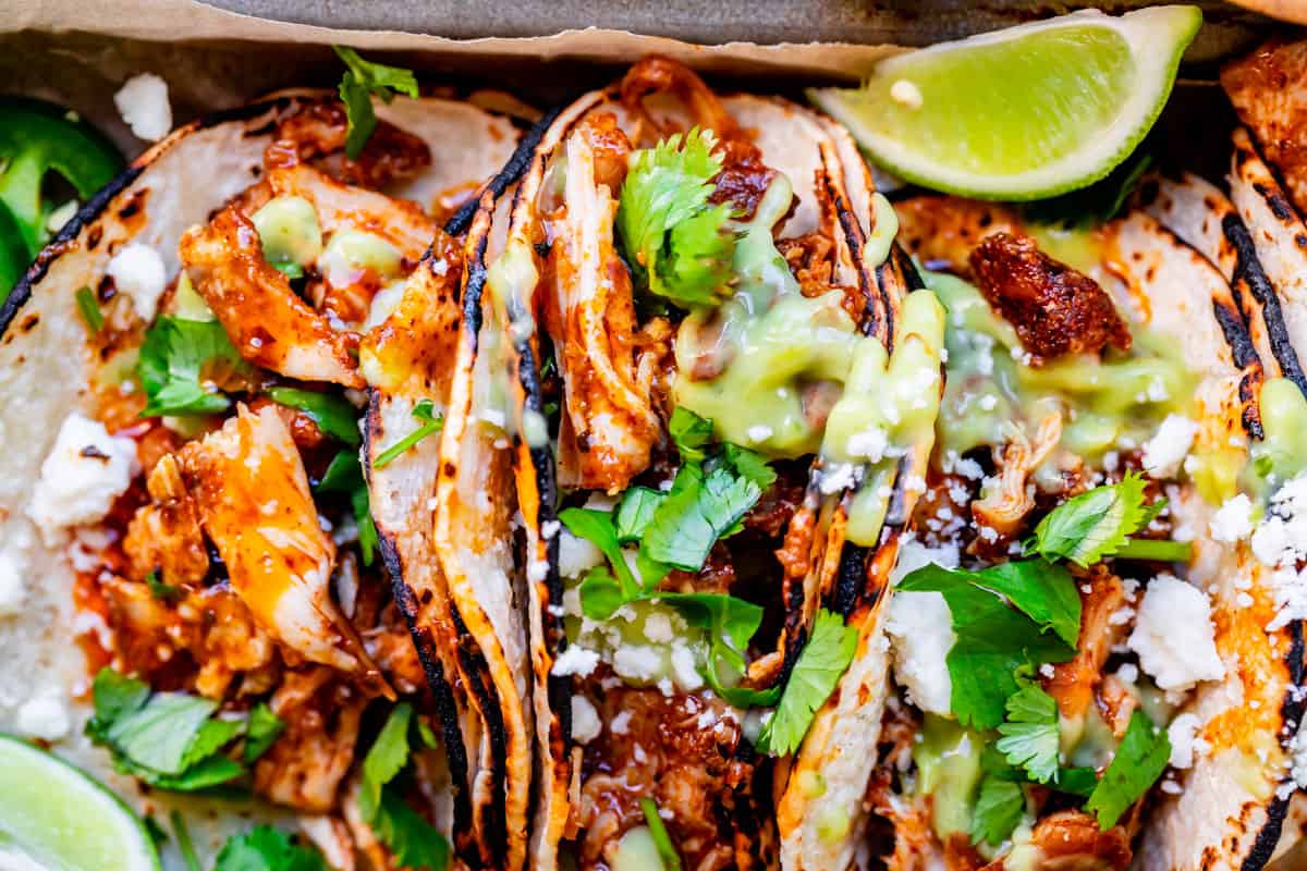 three tacos side by side with charred tortillas, lots of saucy chicken, cilantro, and green salsa.