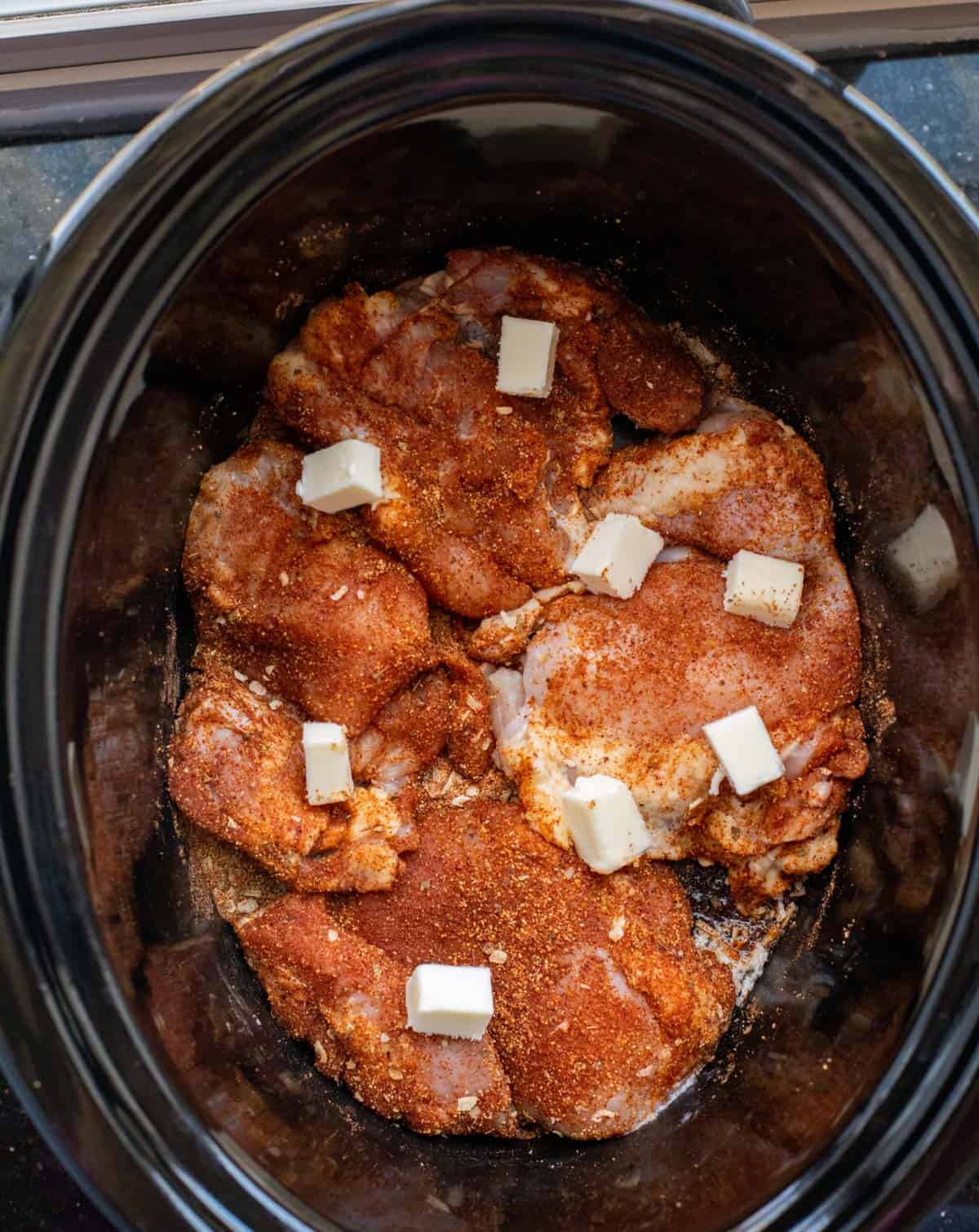 raw chicken thighs covered in a spice dry rub sitting in a crockpot, dotted with butter.