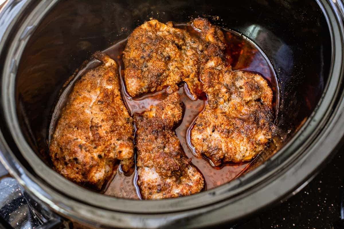 four cooked chicken thighs in a crockpot, covered in seasonings and meat drippings.