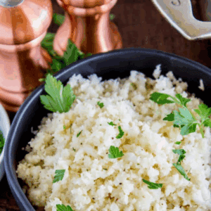 A black bowl filled with cauliflower rice, garnished with parsley, sits on a wooden surface with fresh herbs and copper salt and pepper shakers in the background. Text above reads Cauliflower Rice.