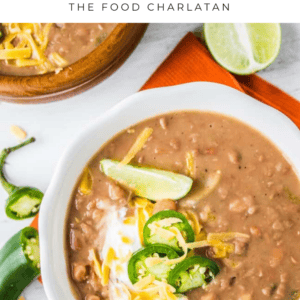 A bowl of Mexican beans pinto soup topped with shredded cheese, sliced jalapeños, sour cream, and lime wedges. A spoon, lime halves, and another bowl sit nearby on a white table with an orange napkin.