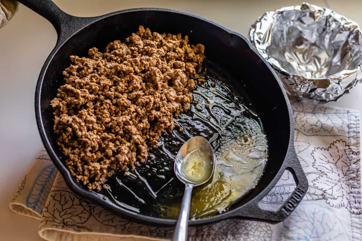 draining grease from a cast iron skillet full of cooked ground beef.