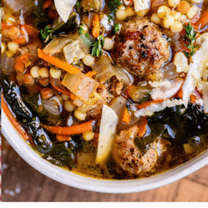 Close-up of a bowl of Italian wedding soup with savory meatballs, pasta, vegetables, and fresh herbs, topped with grated cheese. Text below reads: Seriously delicious Italian Wedding Soup – The Food Charlatan.