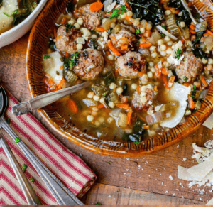 A bowl of Italian wedding soup with savory meatballs, pasta, carrots, spinach, and grated cheese is served with a spoon on a wooden table beside another bowl and a red-striped napkin.