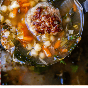 A ladle filled with Italian wedding soup, featuring vegetables, pasta, greens, and a browned meatball, is held above a pot of soup. Text below reads “Italian Wedding Soup – The Food Charlatan.”.