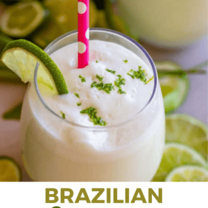 A glass of creamy Brazilian lemonade topped with lime zest, garnished with a lime wedge and a pink polka-dot straw. Sliced limes are scattered in the background. Text below reads, BRAZILIAN Limeade THE FOOD CHARLATAN.