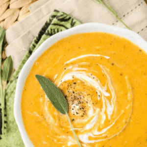 Bowl of creamy roasted butternut squash soup with a swirl of cream, black pepper, and a sage leaf on top, placed on a patterned napkin and woven placemat. Text above reads Roasted Butternut Squash Soup.