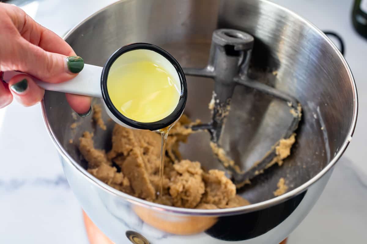adding oil in a measuring spoon to beaten butter and sugar in a stand mixer bowl for cupcake batter.