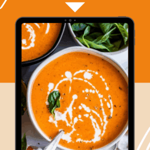 A bowl of creamy tomato soup garnished with a swirl of cream and fresh basil sits on a table, with a spoon beside it. The text reads Creamy Tomato Soup and thefoodcharlatan.com, highlighting a delicious tomato soup recipe.