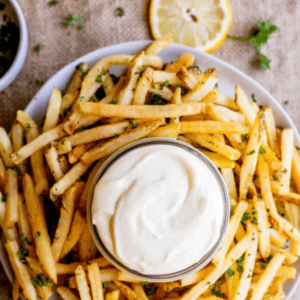A plate of seasoned French fries surrounds a small bowl of creamy garlic aioli, garnished with chopped herbs and lemon slices. Text at the top reads, Garlic Aioli.