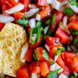 A close-up of fresh pico de gallo with diced tomatoes, onions, cilantro, and jalapeños, featuring a tortilla chip dipped in the vibrant mixture. The text above reads Pico de Gallo.