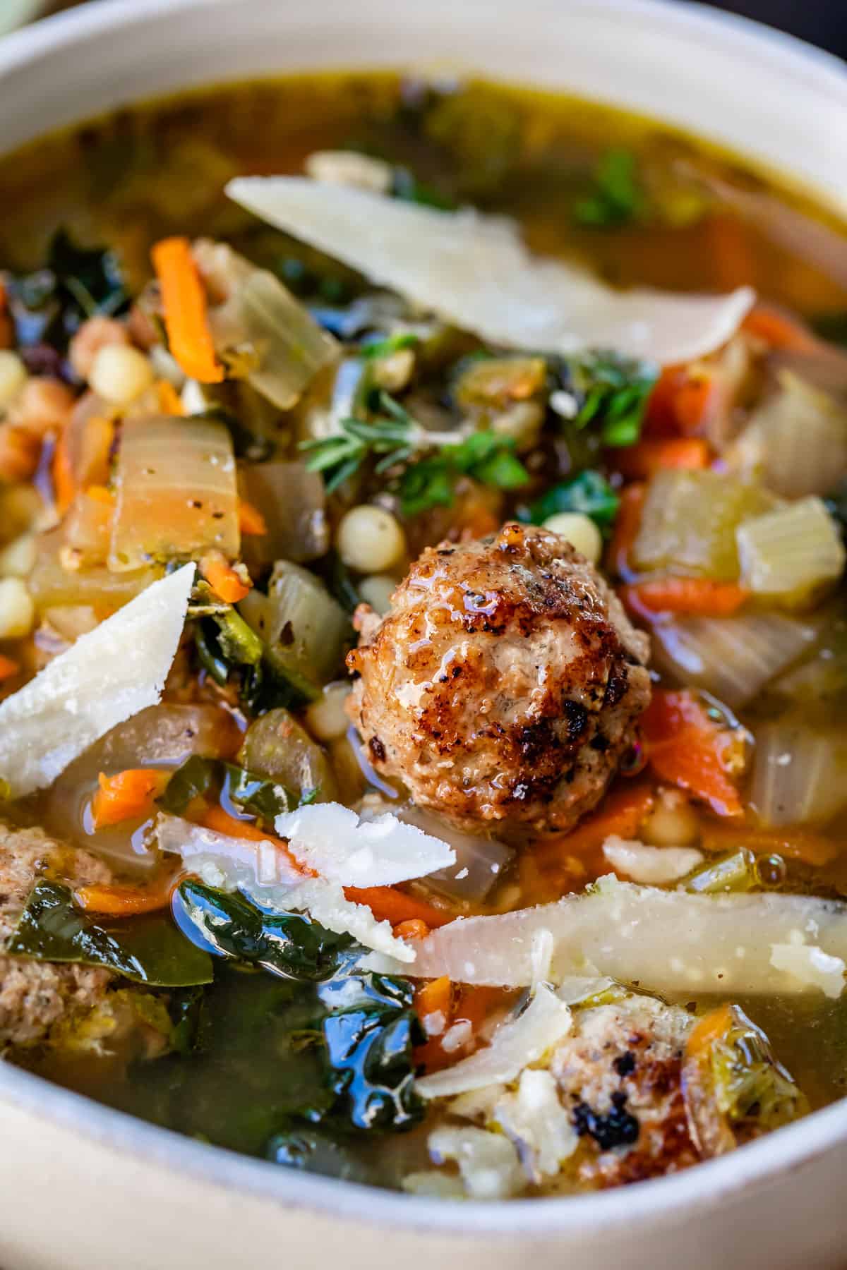 close up featuring a meatball with the broth, greens, and pasta of italian wedding soup behind.