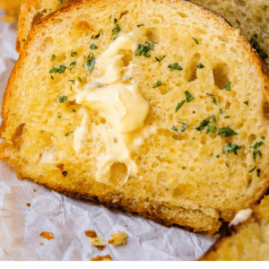 Sliced garlic bread topped with melted butter and fresh chopped herbs, arranged in a row on parchment paper. Text reads: The very finest garlic bread of your life. The Food Charlatan.
