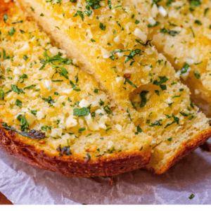 Close-up of two slices of garlic bread topped with chopped parsley and melted butter, featuring a golden, crispy crust. Text at the bottom reads: The Very Best Garlic Bread of Your Life - The Food Charlatan.