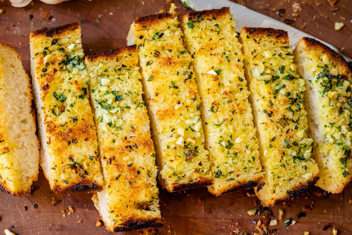 garlic bread made in the oven, sliced and lined up on a cutting board with knife in background.