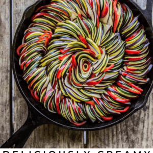 A cast iron skillet filled with neatly arranged, spiral slices of zucchini, eggplant, and red peppers sits on a rustic wooden table next to a bulb of garlic. Text reads: “Deliciously Creamy Roasted Garlic Mediterranean Ratatouille.”.