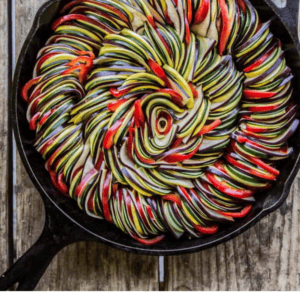 A black cast iron skillet filled with neatly arranged, colorful slices of zucchini, eggplant, and tomato forms a spiral pattern on a rustic wooden surface; fresh vegetables add vibrant appeal while a bulb of garlic sits nearby.