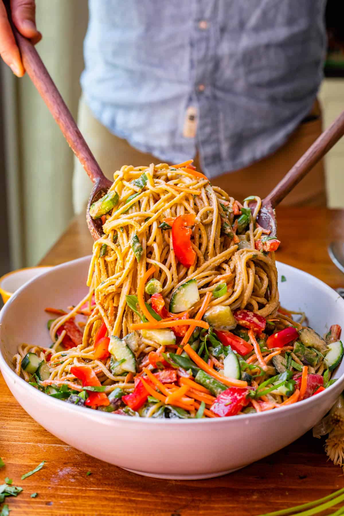 ceramic bowl with sesame noodles with vegetables being lifted up by two wooden salad spoons.