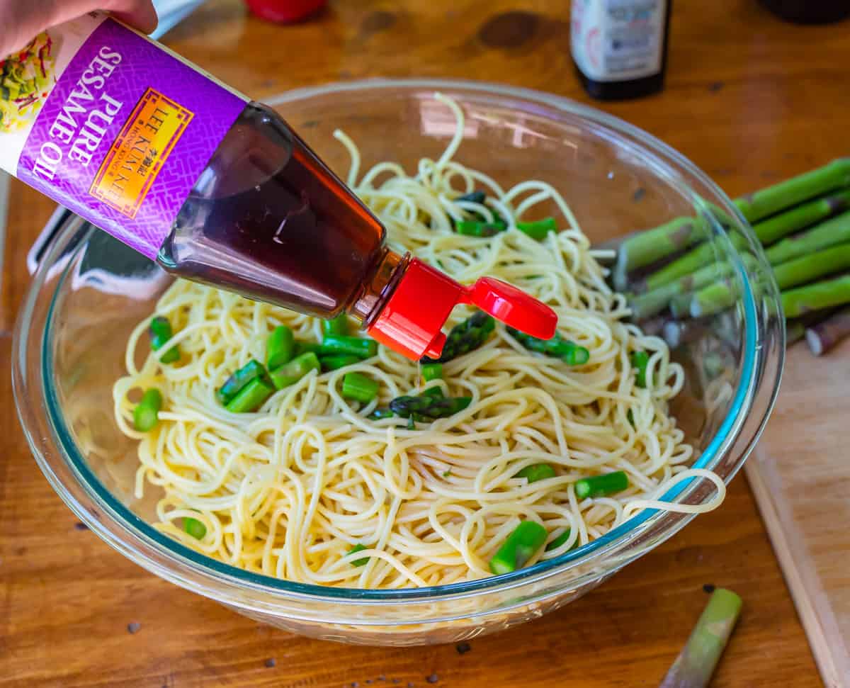 sesame oil being poured from a bottle over cooked noodles and vegetables.