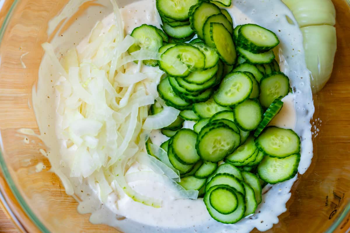 Clear glass bowl with sliced English cucumbers, onions, cream, and spices.