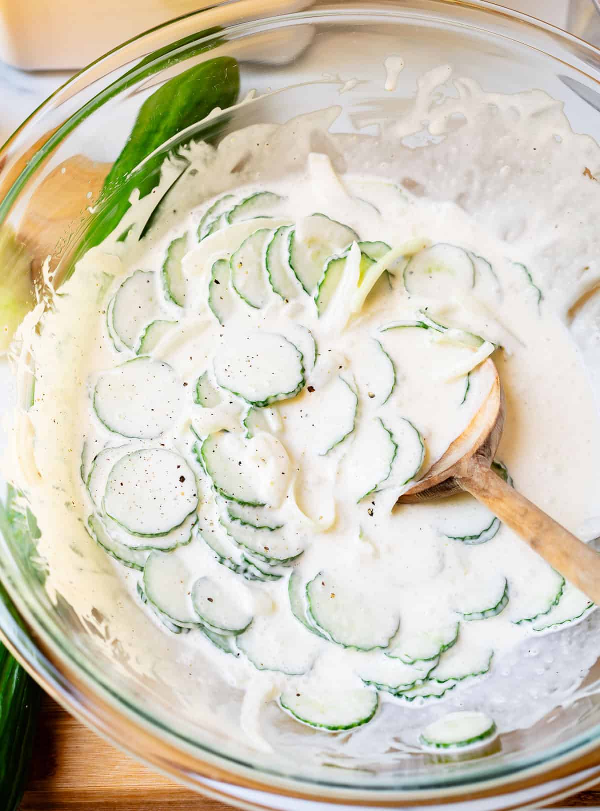 Mixing creamy cucumber salad in large clear glass bowl with a wooden spoon.