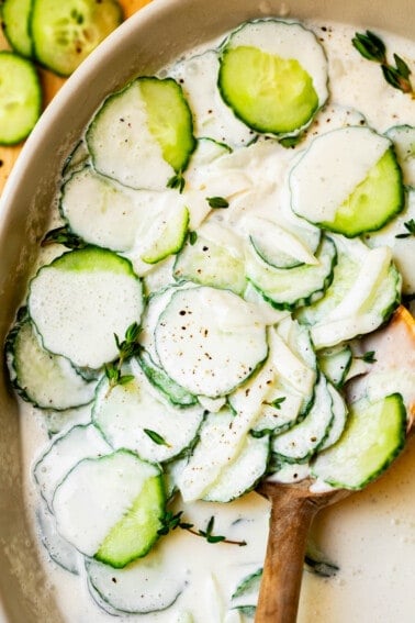 creamy cucumber salad with onions and thyme in a white bowl with wooden spoon.
