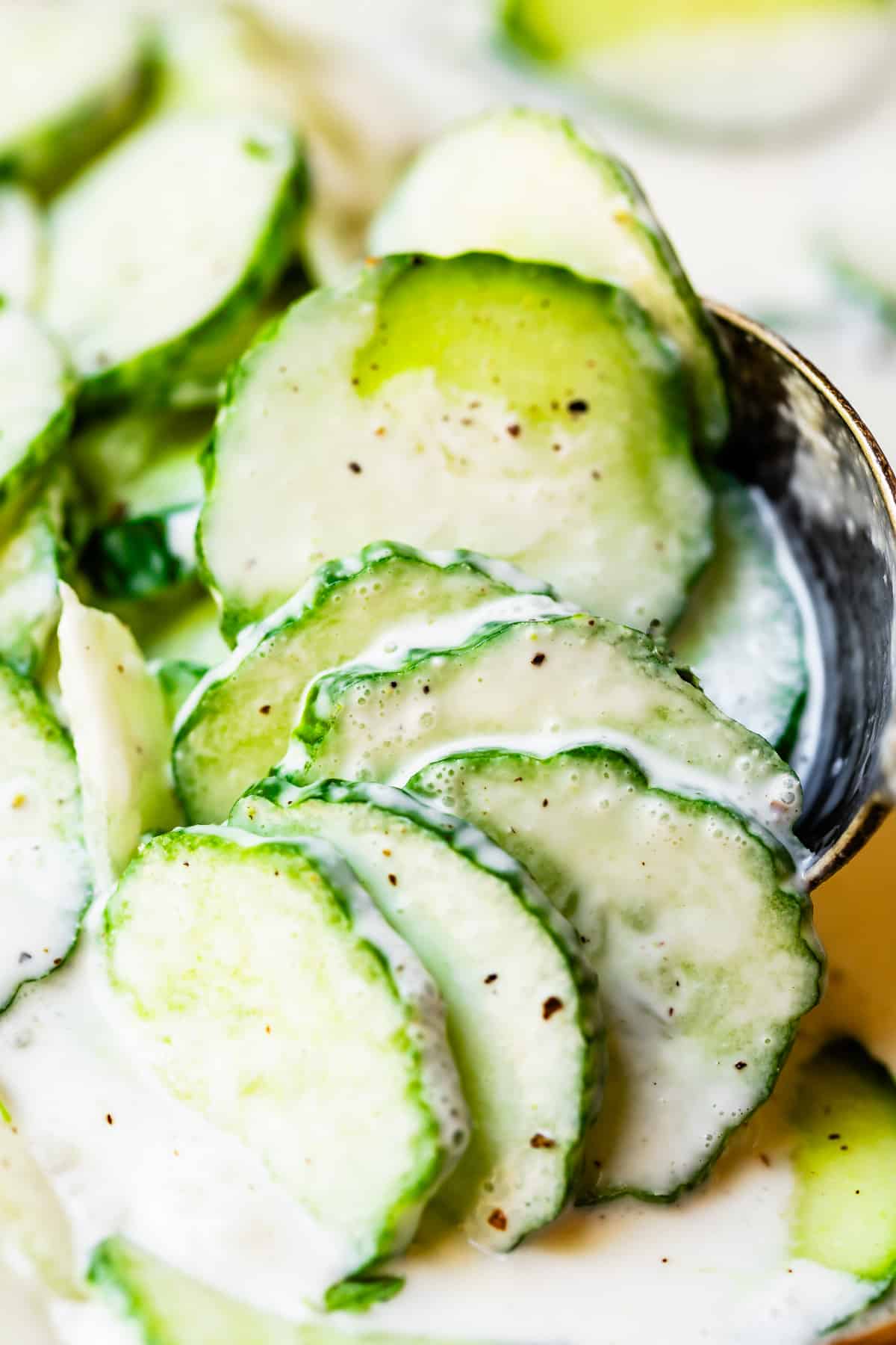 close up shot of cucumbers in a creamy sauce with pepper, held up by metal spoon.