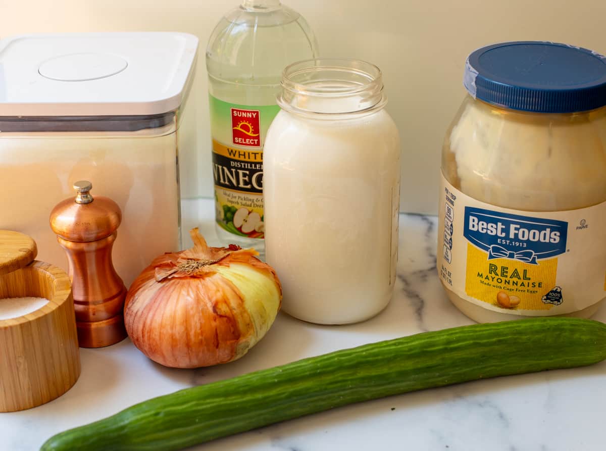 Picture of ingredients laid out for cucumber salad including mayonnaise, cream, onion, English cucumber, vinegar, salt and pepper, and sugar.