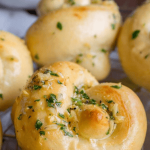 Close-up of golden, freshly baked Garlic Knots topped with chopped parsley and garlic butter, with text overlay: The Ultimate Garlic Knots From Scratch. Logo in corner reads The Food Charlatan.