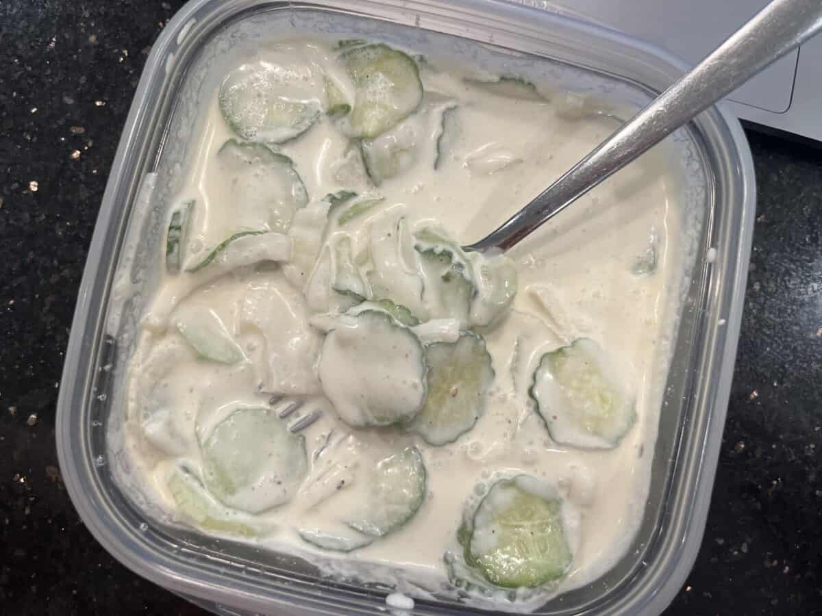 overhead shot of tupperware full of creamy cucumber salad with fork sticking out, on a black counter. 