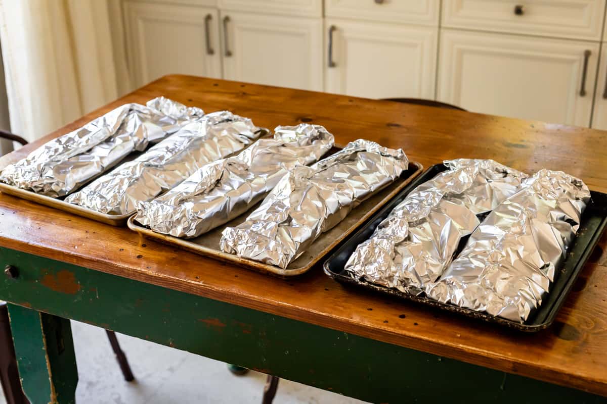 6 racks of ribs wrapped in foil on baking sheets, laid out on a kitchen table.