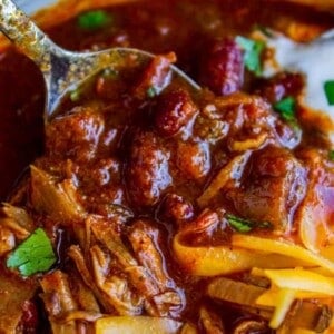 A close-up of a spoonful of hearty chili loaded with chunks of meat, beans, and tomato in a thick, red sauce, topped with melted shredded cheese and fresh cilantro.