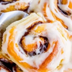 A close-up of freshly baked cinnamon rolls topped with creamy vanilla icing, with one roll being lifted from the tray by a metal spatula. The rolls appear soft, golden brown, and gooey.